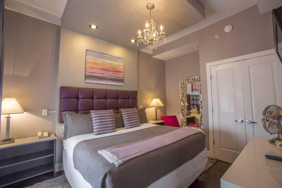 A bedroom with a large bed, two bedside lamps, a chandelier, a door, and a mirror with a decorative frame.