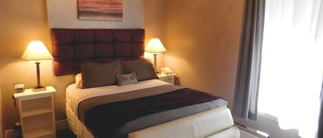 Habitación de hotel con cama, dos lámparas de noche, mesita de noche, una silla, candelabro, un cuadro en la pared y ventana con cortinas.