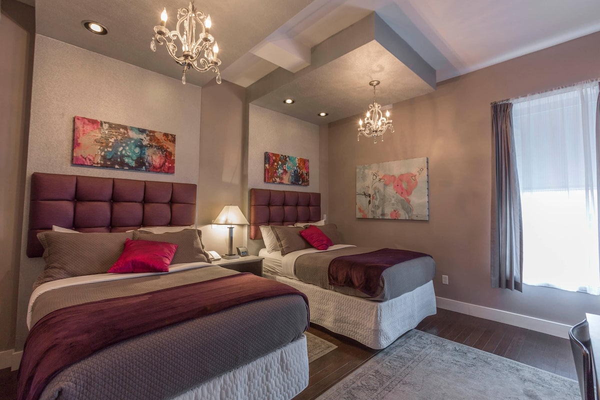 A hotel room with two beds, a chandelier, artwork on the walls, and a window with curtains.