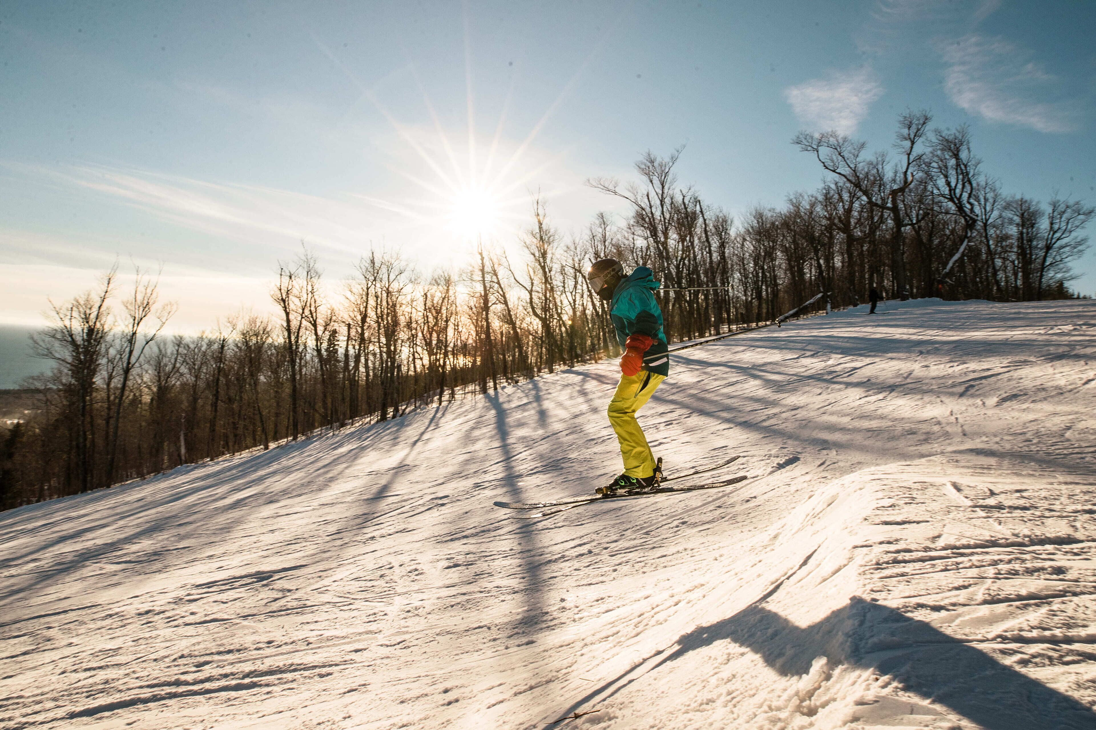 A person skiing down a snowy slope with trees in the background and the sun shining.