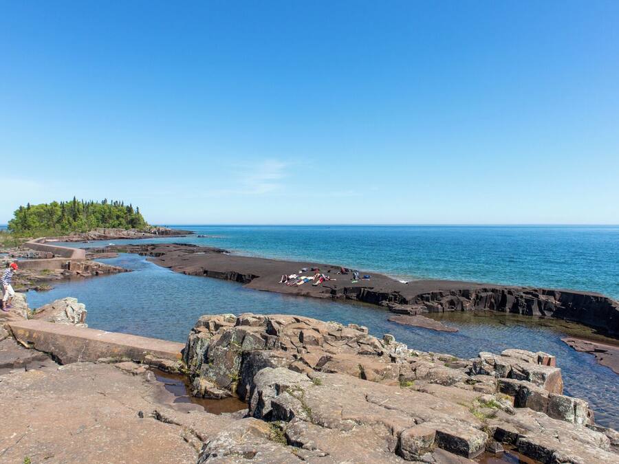 A rocky coastal area with clear blue water, a small island with trees, and people relaxing on the shore.