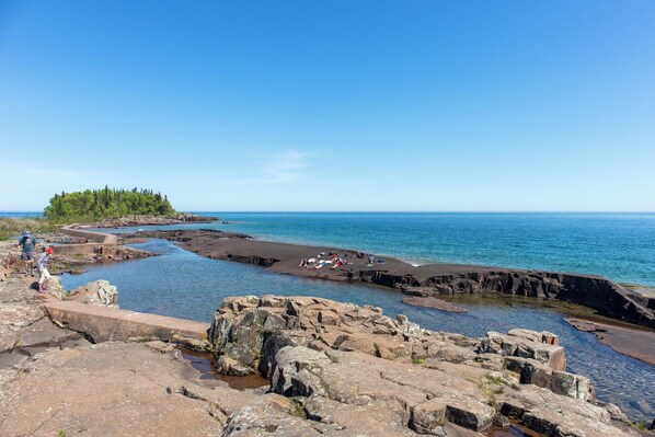 A rocky coastal area with clear blue water, a small island with trees, and people relaxing on the shore.