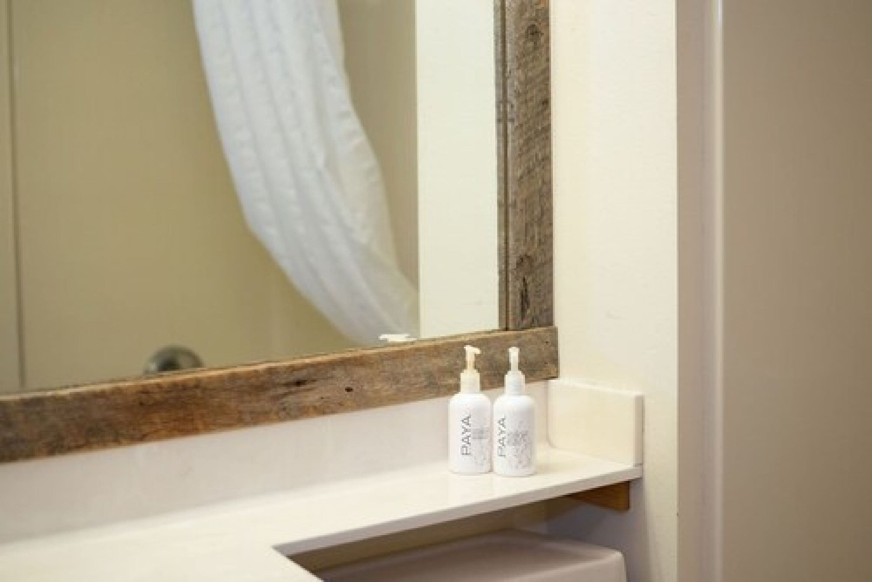 Une salle de bain avec un lavabo blanc, un miroir à cadre en bois et deux bouteilles de savon Paya posées sur le lavabo.