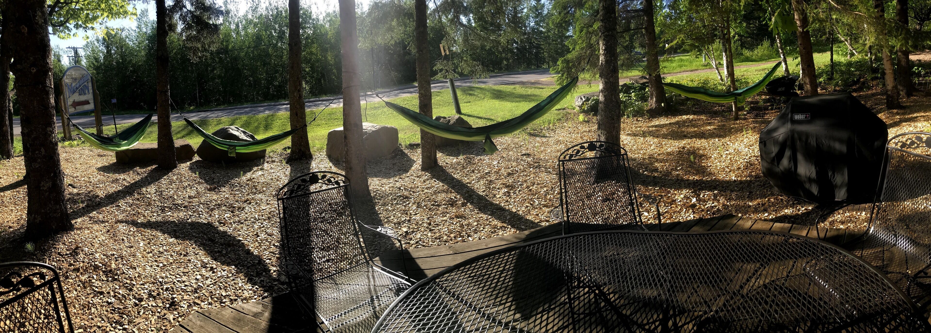A hammock and metal chairs in a wooded area with a road in the background.