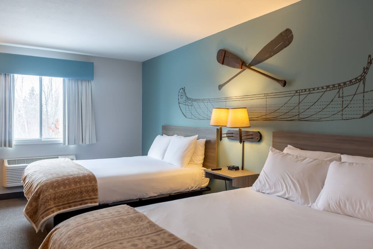 A hotel room with two beds, a window with curtains, a wall-mounted lamp, and a canoe design on the wall.