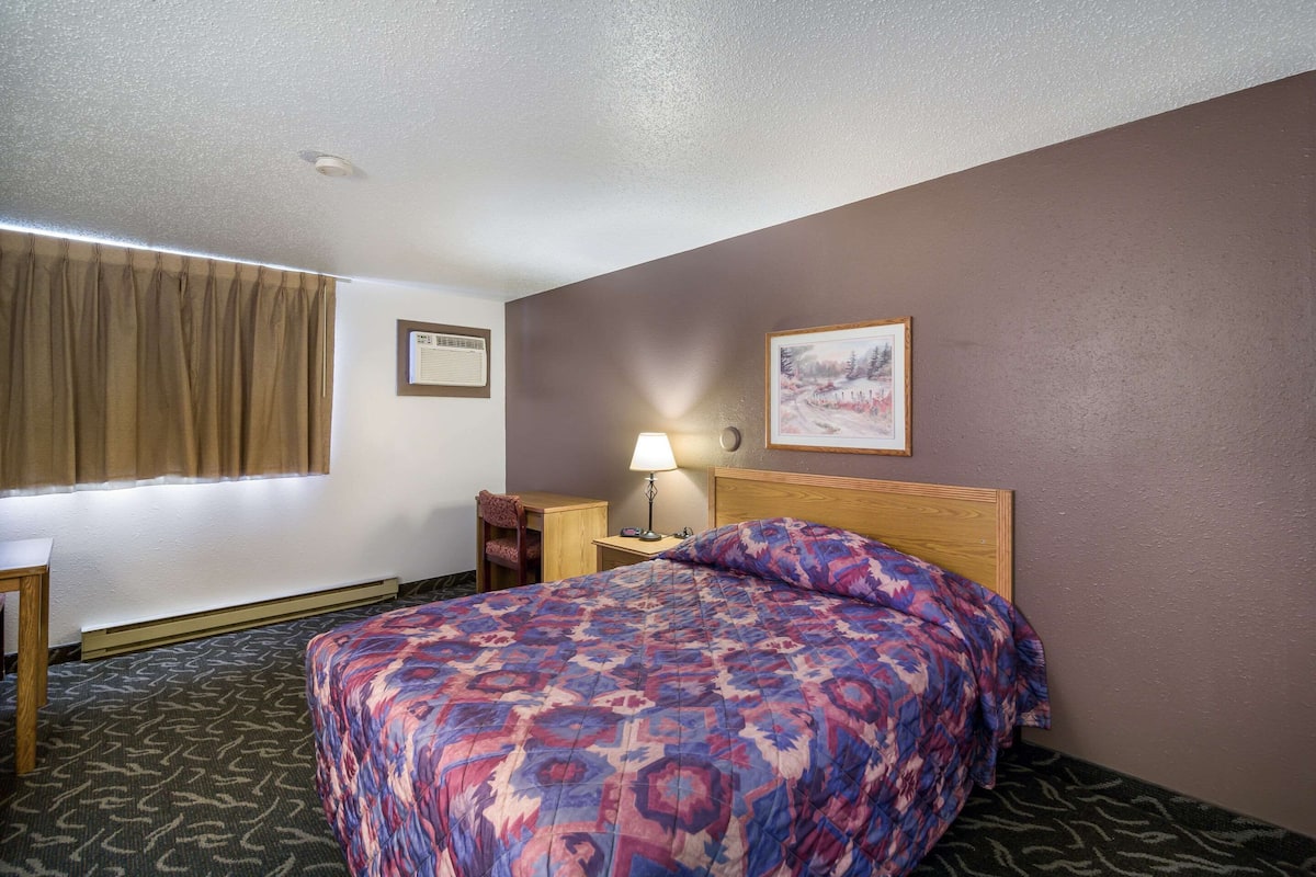 A hotel room with a bed, a nightstand, a lamp, a chair, a window with curtains, and a painting on the wall.