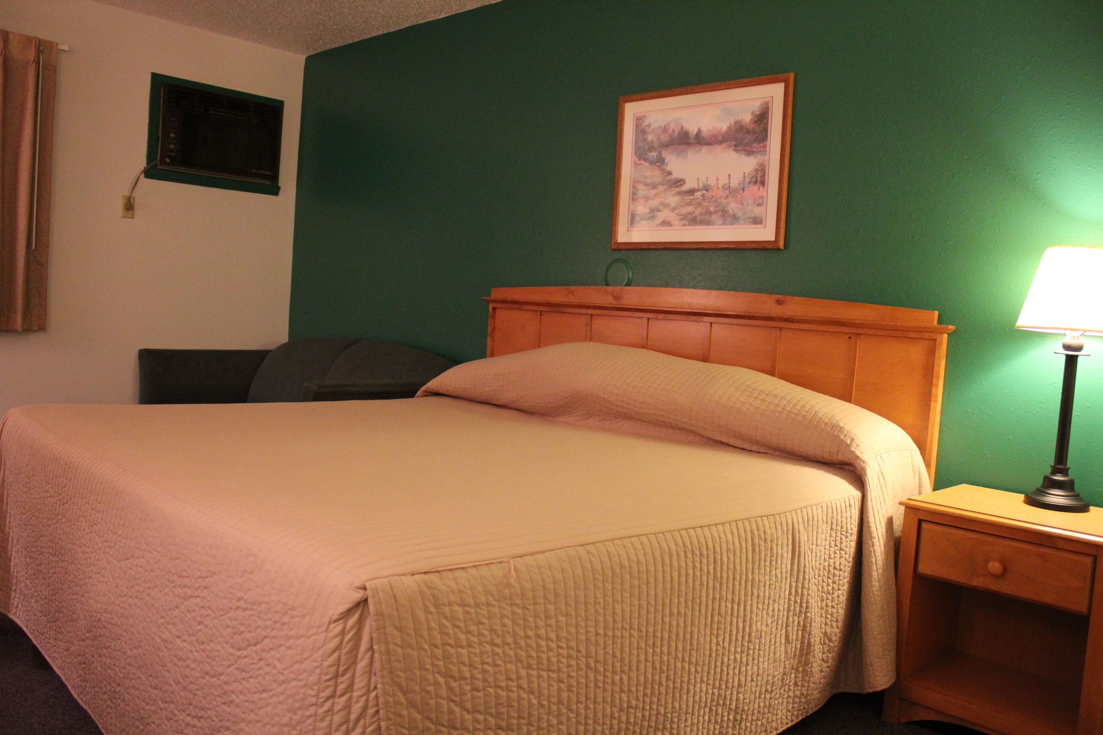 A hotel room with a large bed, a nightstand with a lamp, a painting on the wall, and a window with curtains.