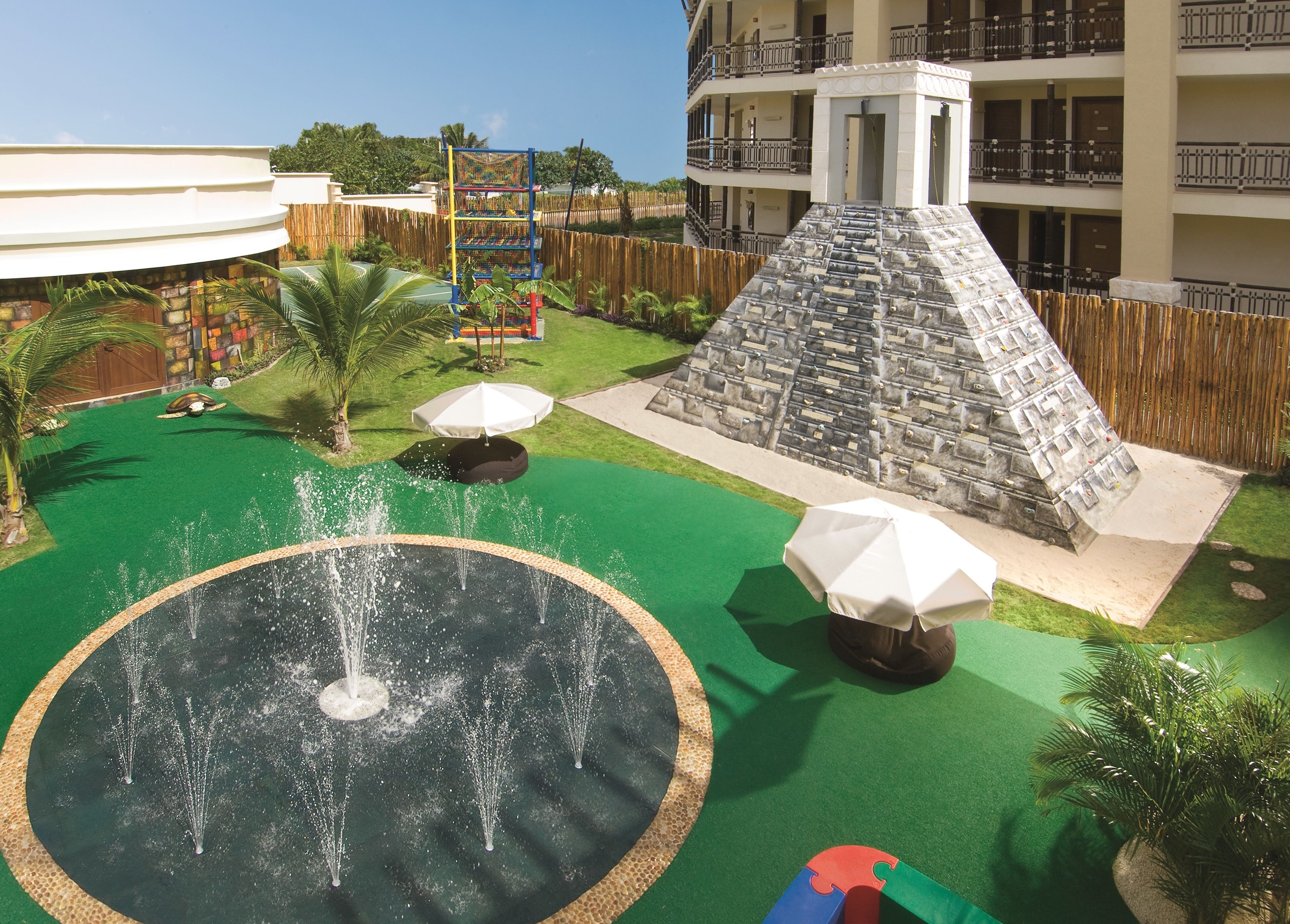 Zona de juegos infantiles con una fuente, palmeras y un edificio de varios niveles al fondo.