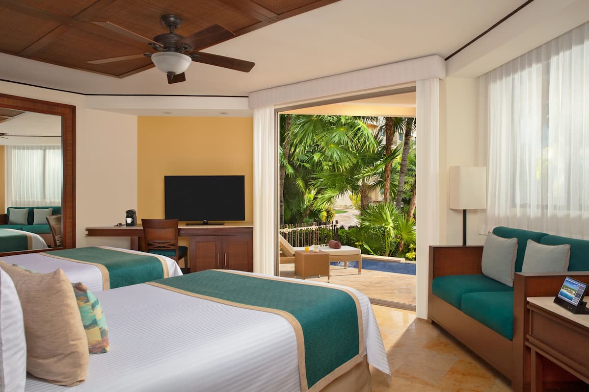 A hotel room with two beds, a TV, a desk, and a balcony with a pool and tropical plants.