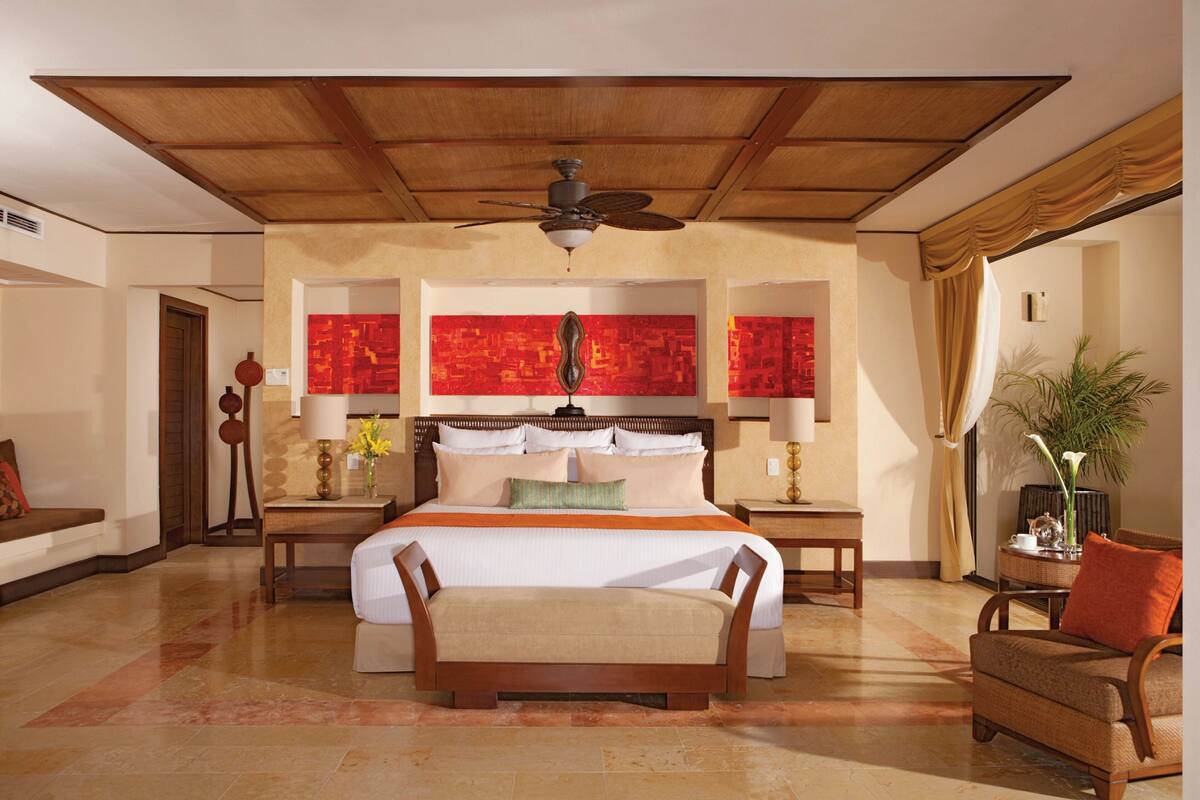 A spacious hotel room with a large bed, a ceiling fan, a nightstand with a lamp, a chair, and a potted plant.