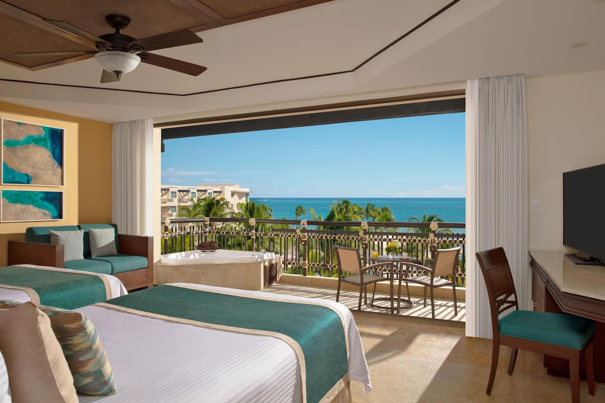 A hotel room with two beds, a balcony with a view of the ocean, a ceiling fan, and a TV.