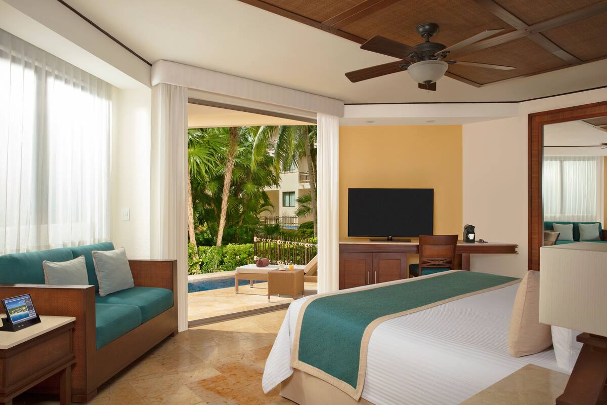 A hotel room with a bed, a TV, a desk, a sofa, and a balcony with a view of palm trees.