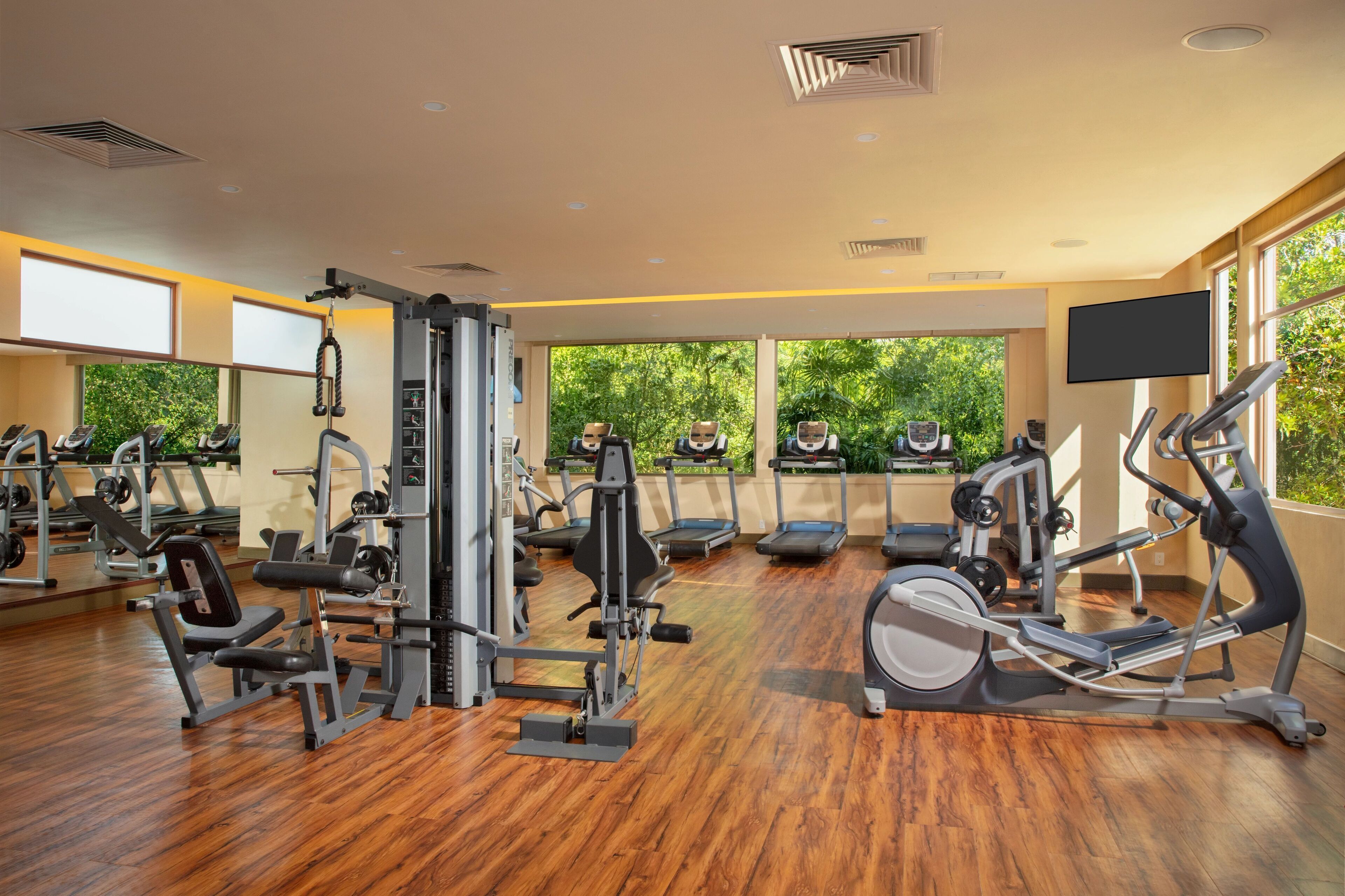Un gimnasio con diversos equipos de ejercicio, ventanas amplias y un televisor en la pared.