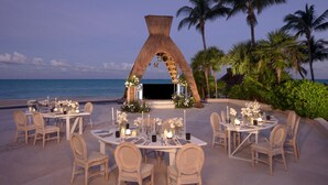 An outdoor wedding setup with tables and chairs arranged on the beach, a decorative archway, and palm trees in the background. - Dreams Riviera Cancun Resort & Spa - All Inclusive (Puerto Morelos)