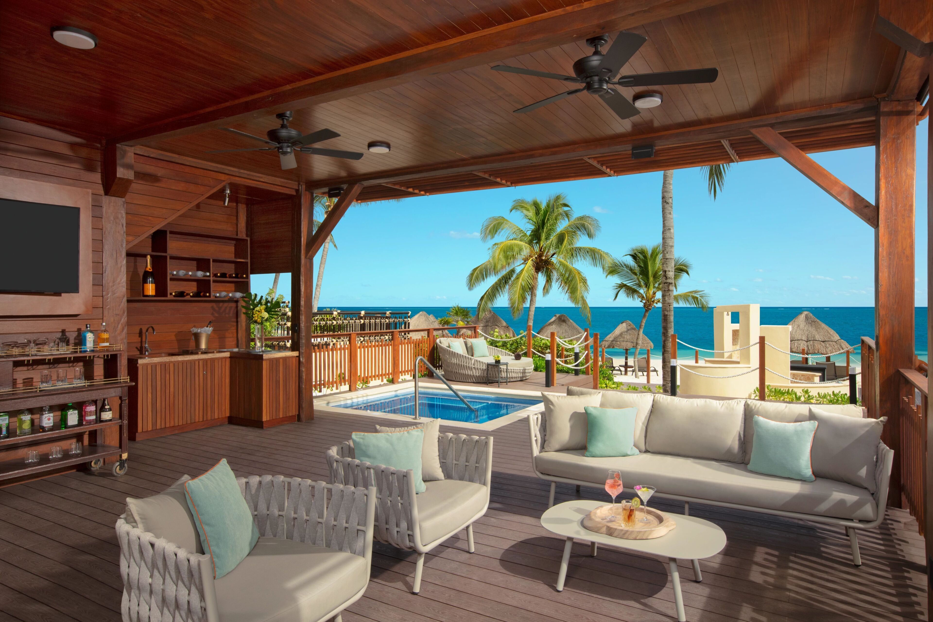 Une terrasse en bois avec une piscine, des chaises longues, un canapé et un coin bar doté d’étagères et de bouteilles.