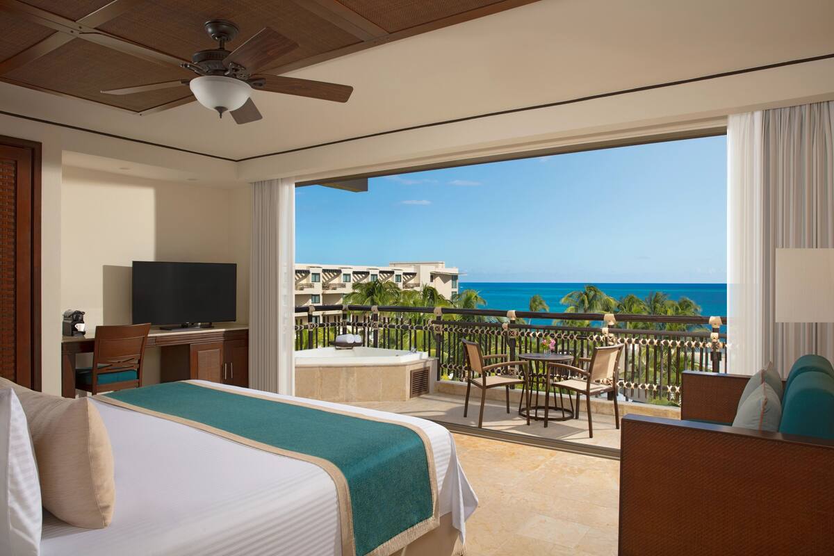 A hotel room with a balcony, a bed, a desk with a TV, and a view of the ocean.