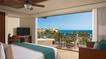 A hotel room with a balcony, a bed, a desk with a TV, and a view of the ocean.