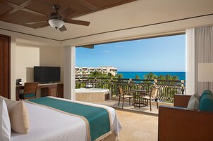 A hotel room with a balcony, a bed, a desk with a TV, and a view of the ocean.