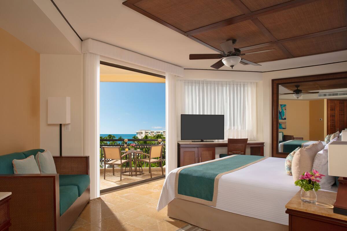 A hotel room with a bed, a TV, a desk, a chair, and a balcony with a view of the ocean.