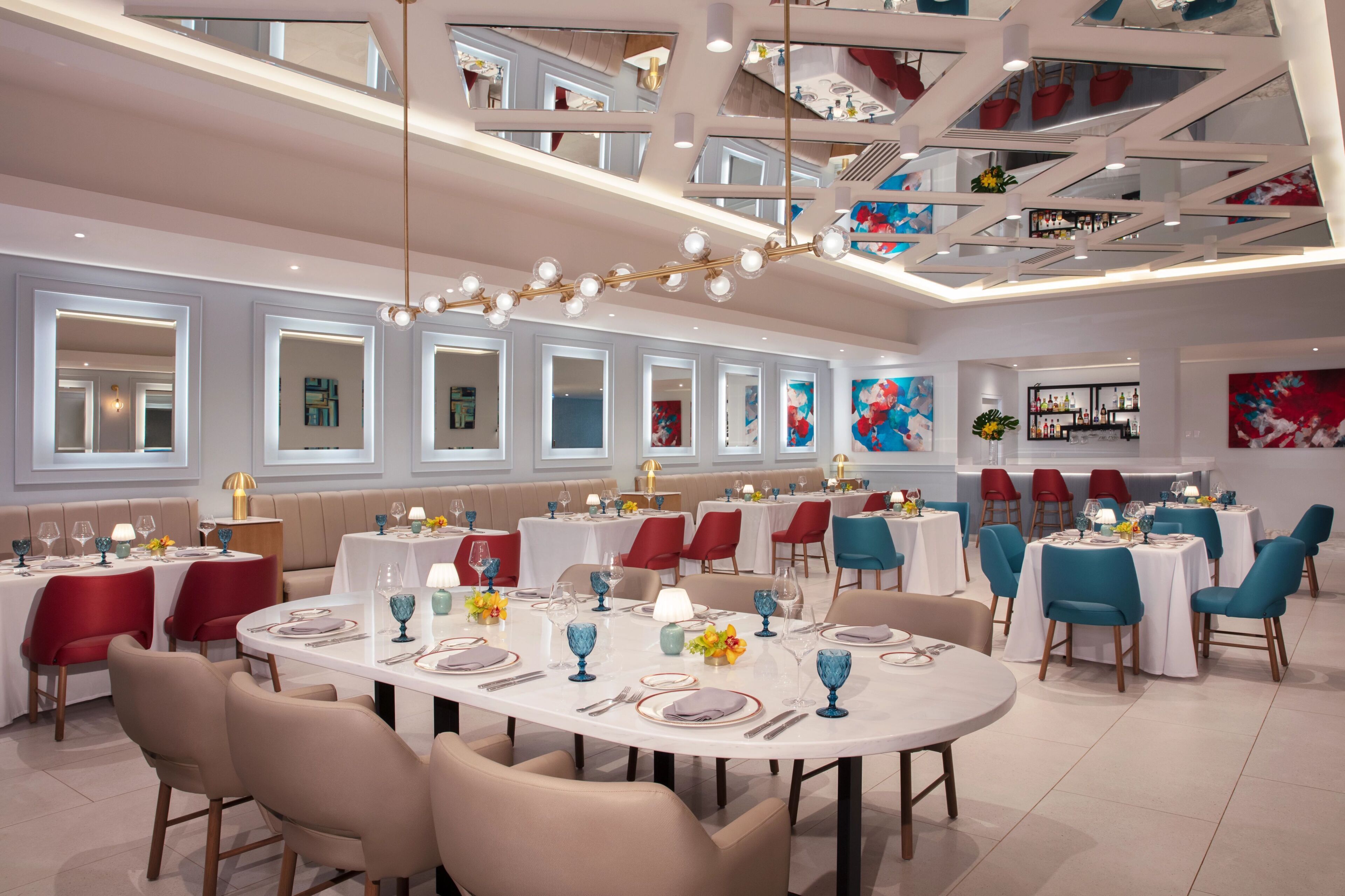 Uma sala de jantar moderna com mesas redondas, cadeiras vermelhas e azuis, e teto alto com design geométrico.