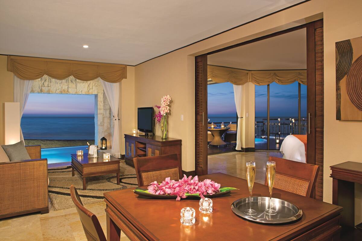 A hotel room with a dining area, a TV, a fireplace, and a view of the ocean.