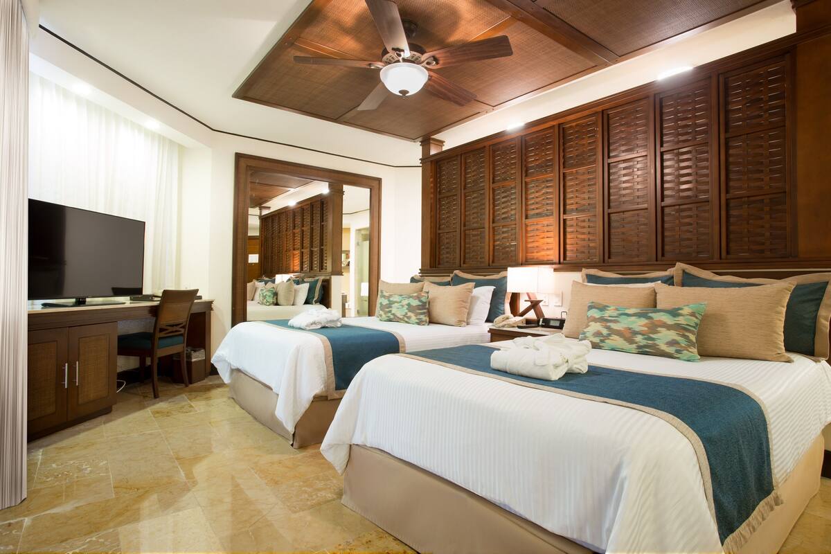 A hotel room with two beds, a desk, a television, and a ceiling fan.