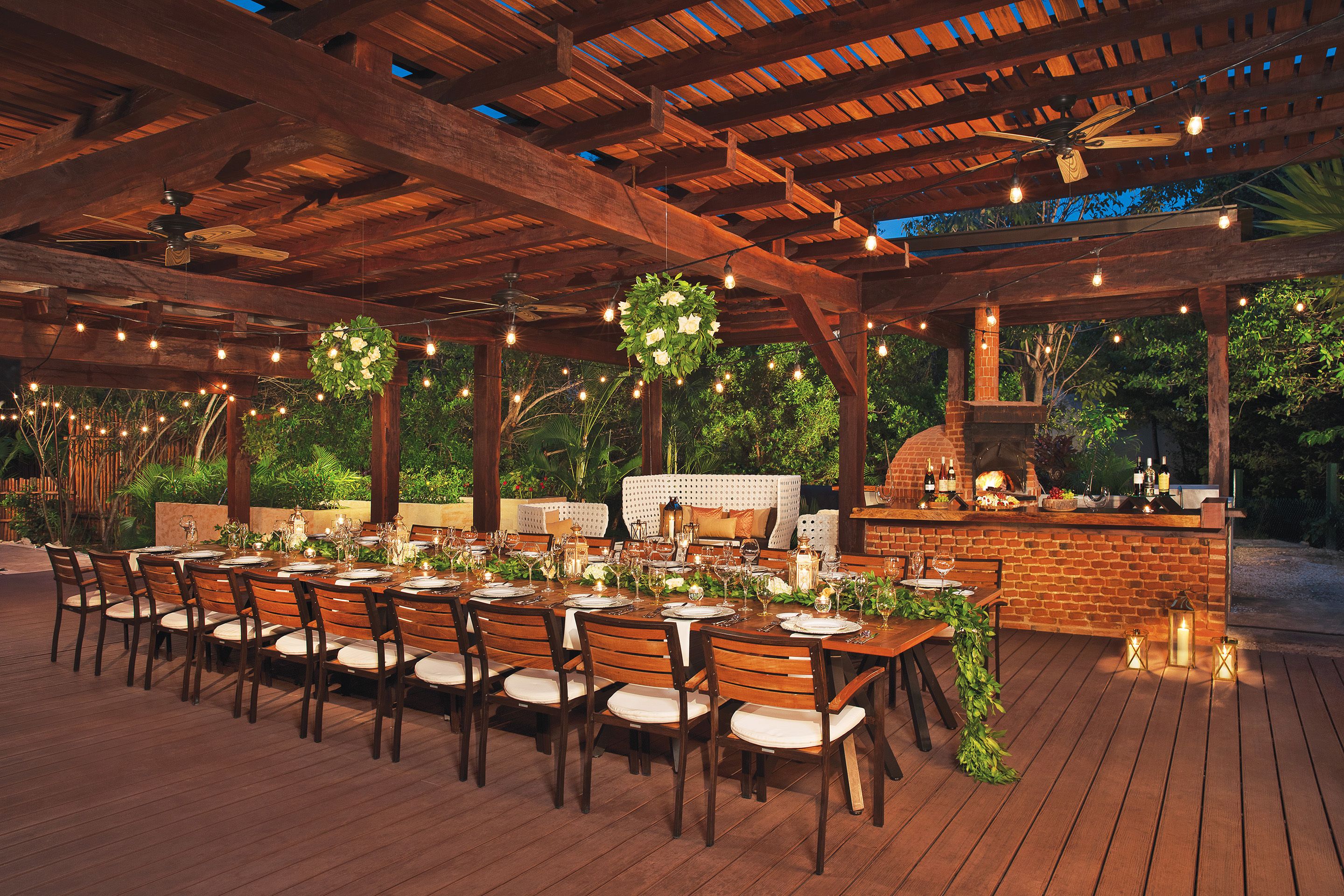 An outdoor dining area with wooden tables and chairs, a brick barbecue, and string lights.