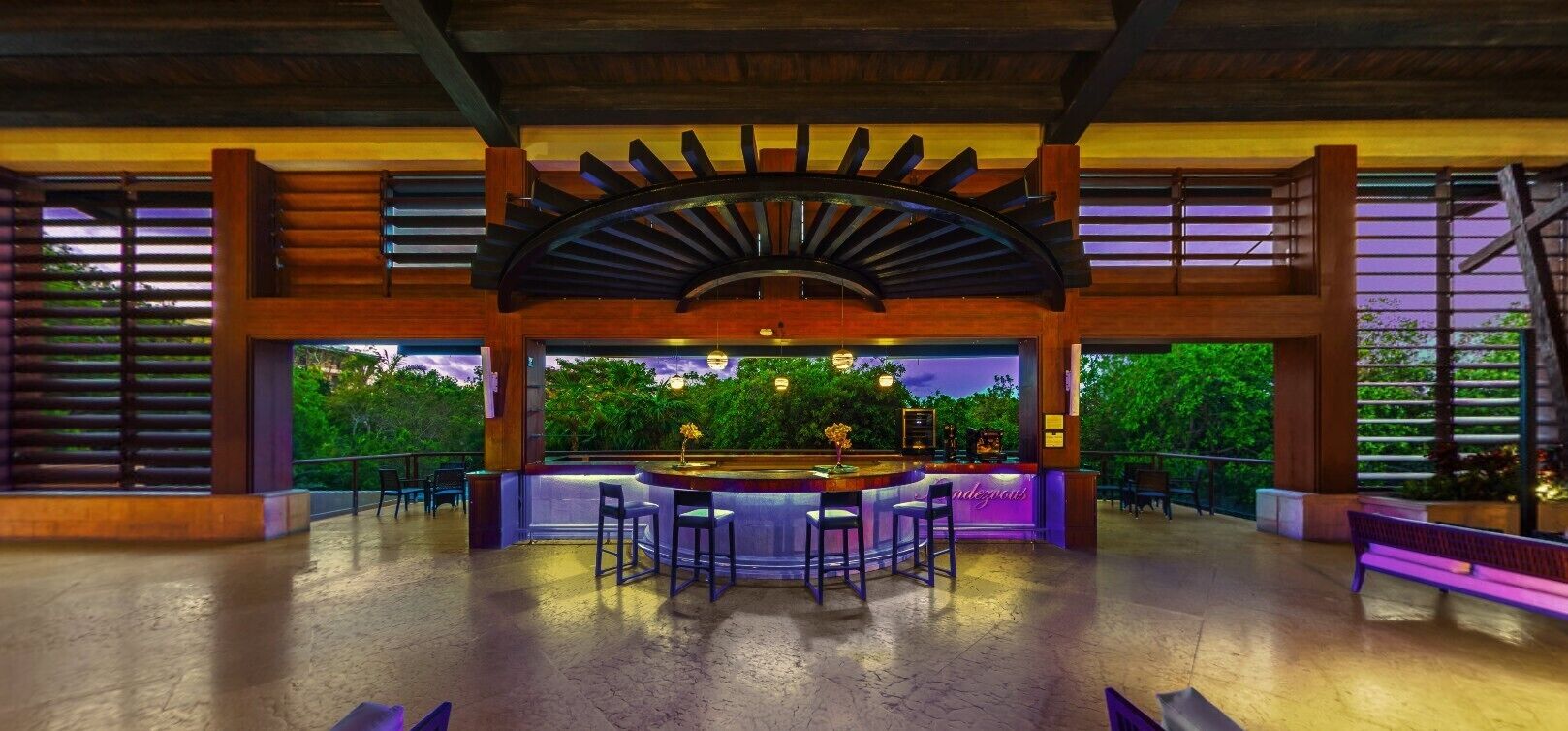 Un área de bar al aire libre con barra central, asientos y vistas a la vegetación.