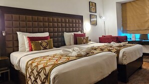 Ein Hotelzimmer mit einem großen Bett, einem kleineren Bett, einem Nachttisch, einer Lampe und einem Fenster mit Jalousien.