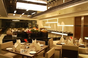 Un espace repas moderne avec des tables dressées pour le service, un buffet proposant des fruits et des légumes, et un bar au design minimaliste.