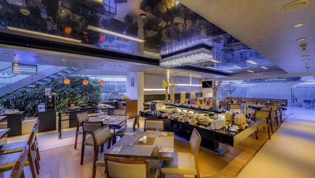 Ein modernes Restaurant mit einer zentralen Bar, Holztischen und -stühlen, umgeben von Pflanzen und Blick auf die Stadt.