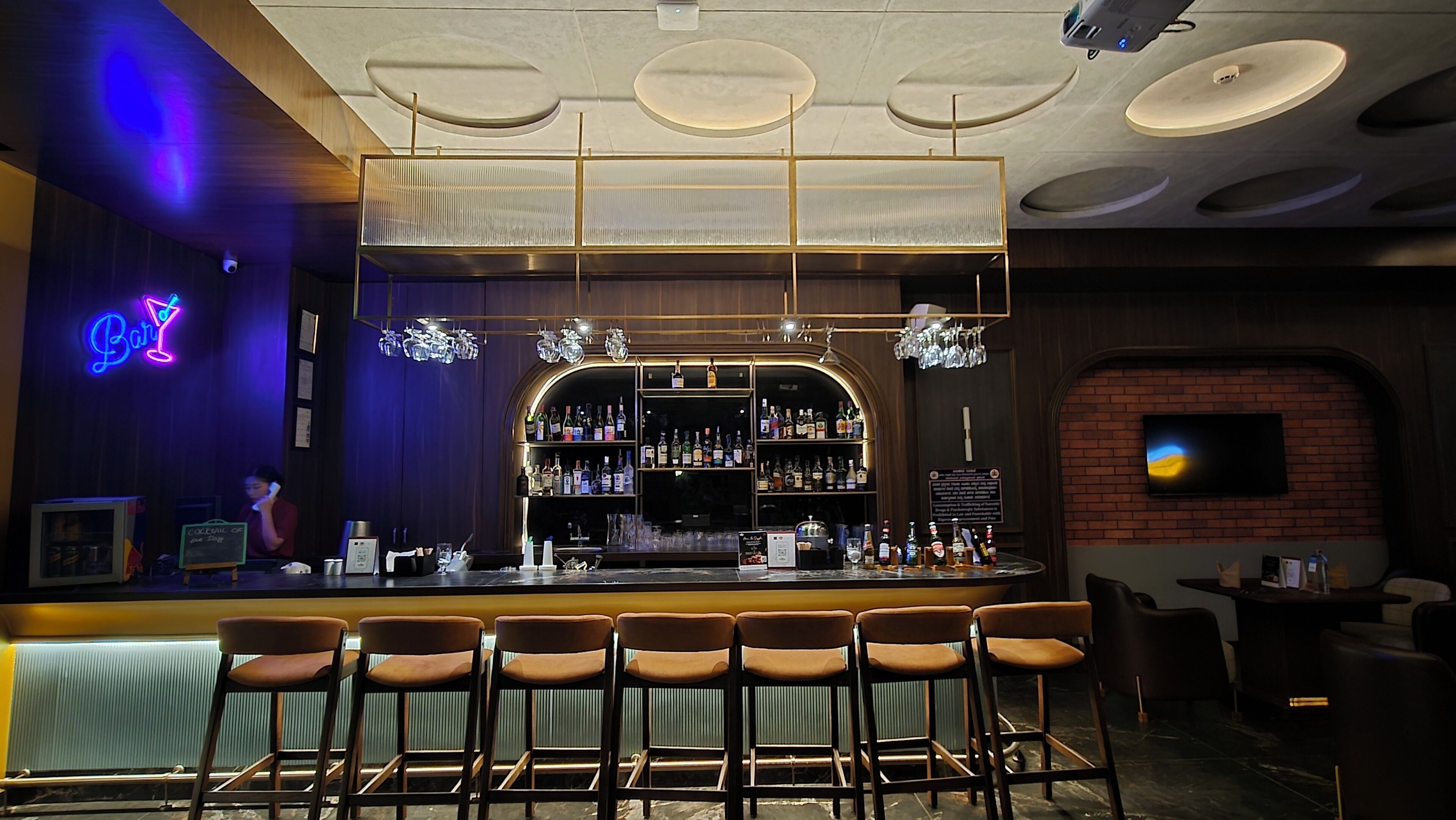 A bar area with a neon sign, shelves of liquor bottles, and a brick wall.