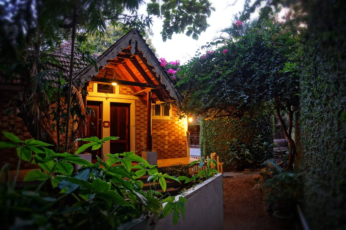 A small, rustic wooden house with a brick wall and a wooden door, surrounded by lush greenery and a pathway leading to it.