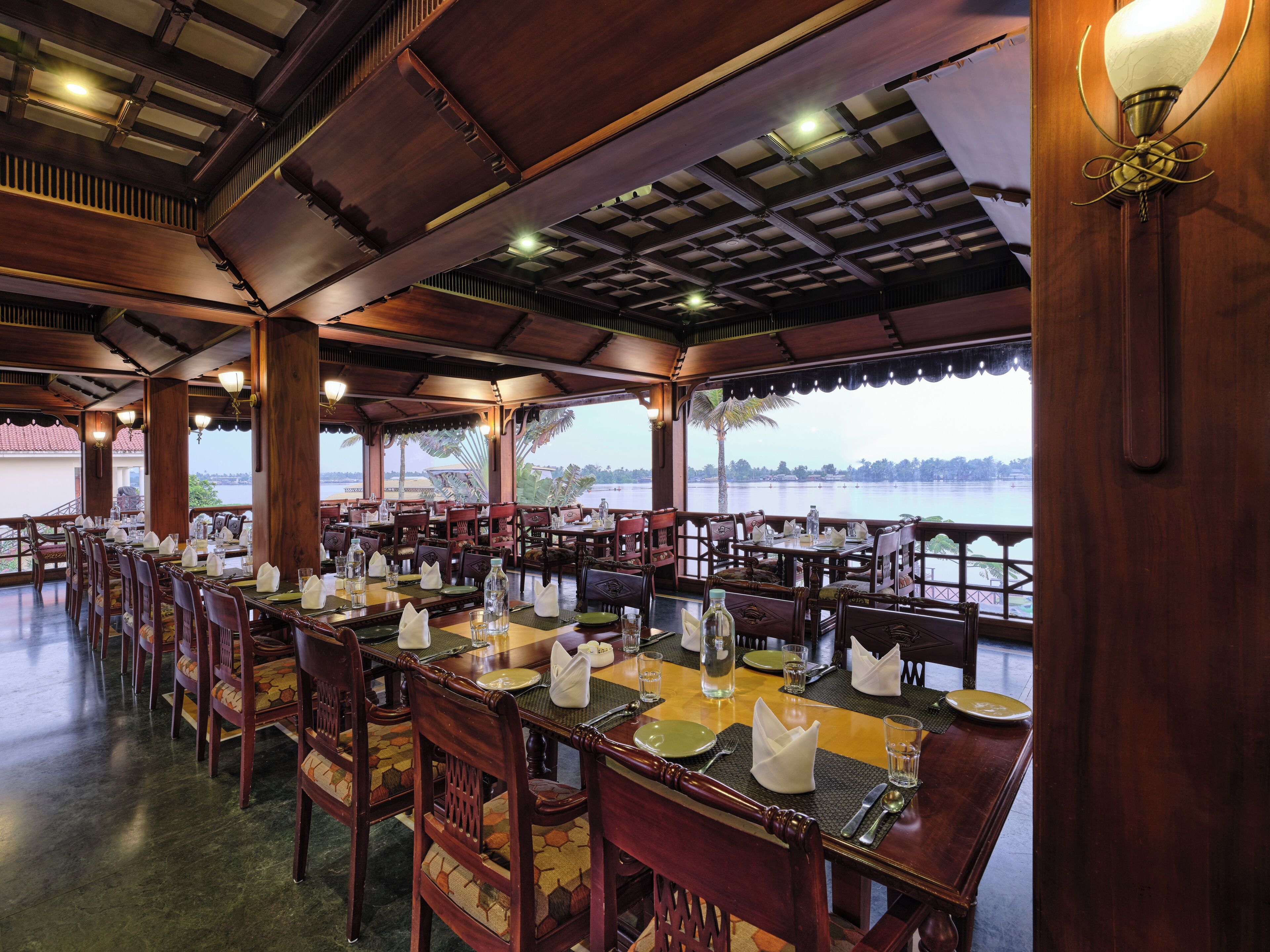 A restaurant with wooden interiors and tables set for dining, overlooking a body of water.