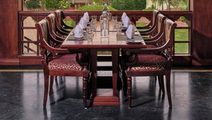 A dining area with a table set for four, surrounded by wooden chairs with red cushions, a view of tropical greenery and palm trees outside. - Sterling Lake Palace Alleppey (Ambalapuzha)