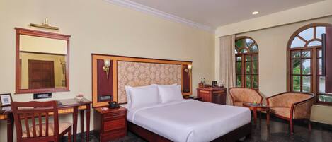 A hotel room with a large bed, a desk, a chair, a nightstand, a mirror, and a window with a view of greenery.