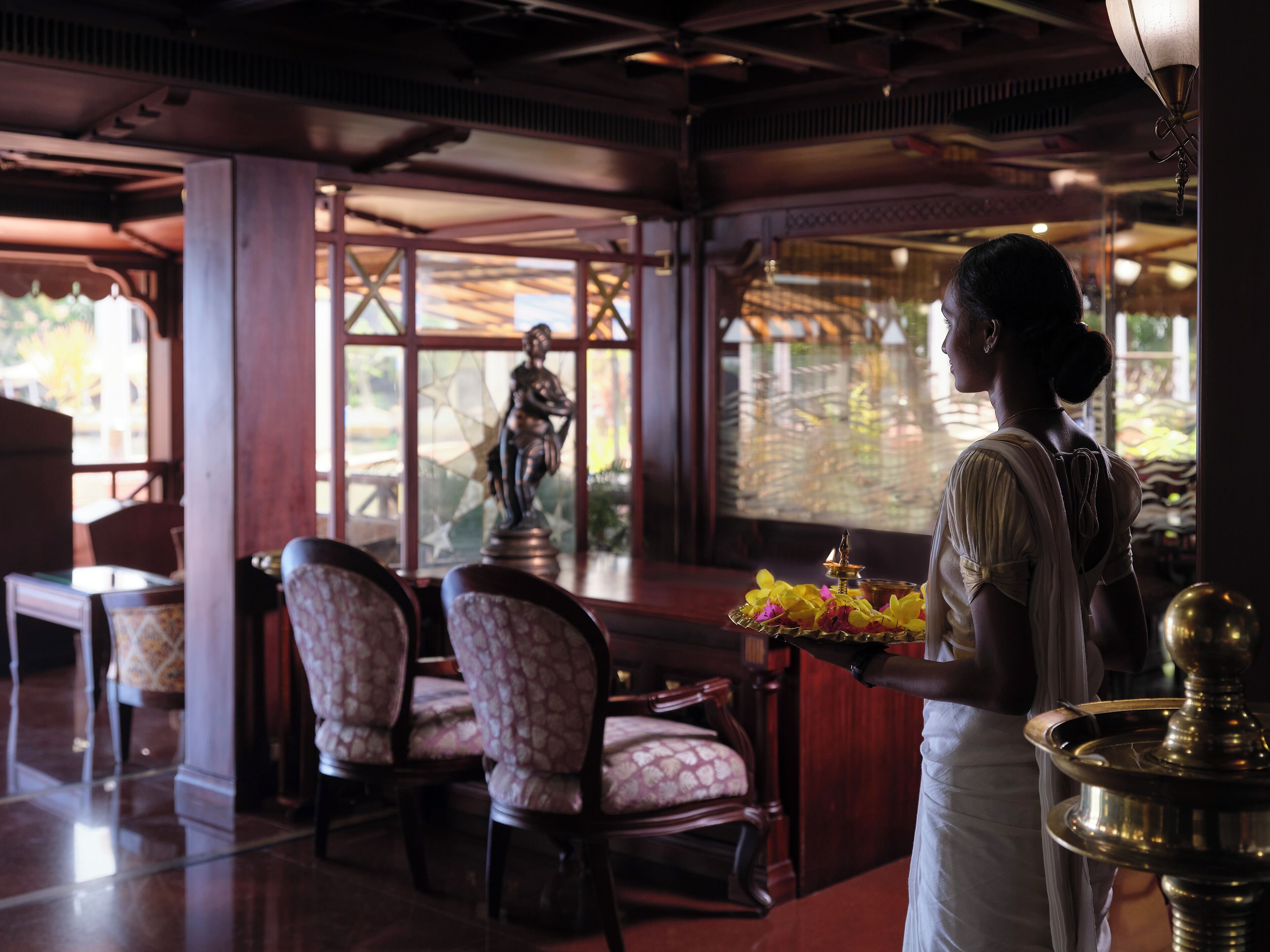 Una persona vestida con traje tradicional sostiene una bandeja con flores y una lámpara, de pie en una habitación con sillas estampadas, una estatua y vistas por la ventana.