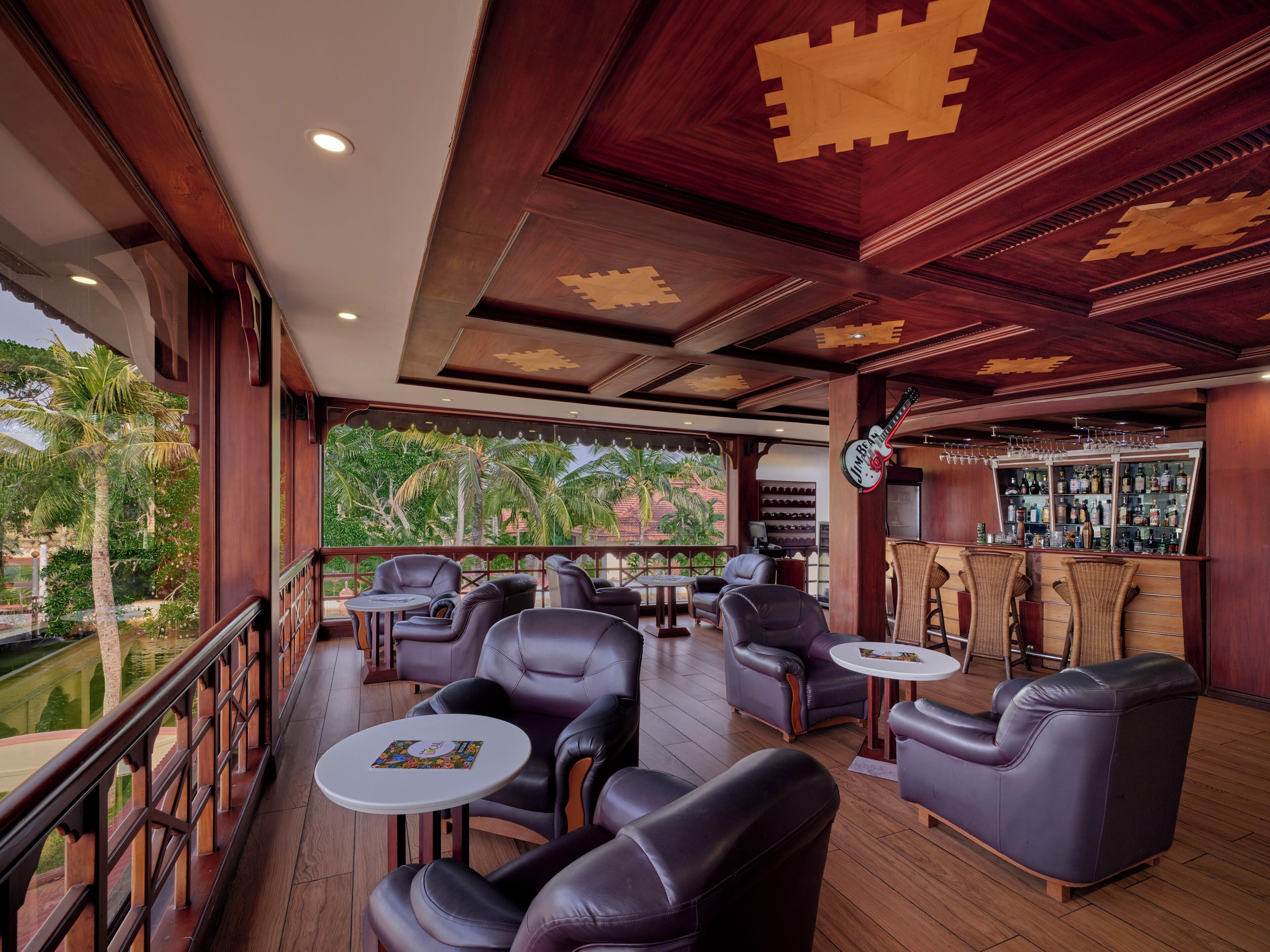 A spacious lounge area with leather chairs and tables, a bar with a guitar decoration, and a view of palm trees through large windows.
