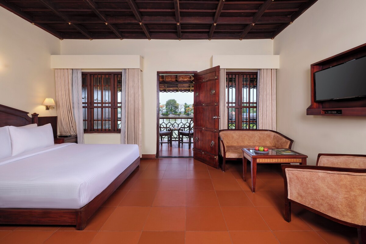 A hotel room with a large bed, a TV mounted on the wall, a seating area with a sofa and chairs, and a balcony with a view.