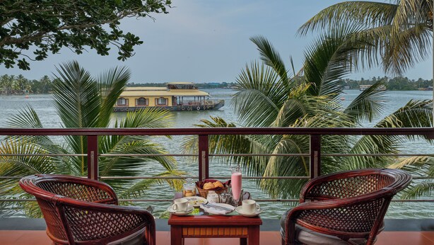 A balcony with two wicker chairs, a small table with a drink and snacks, and a view of a houseboat on the water. - Sterling Lake Palace Alleppey (Ambalapuzha)