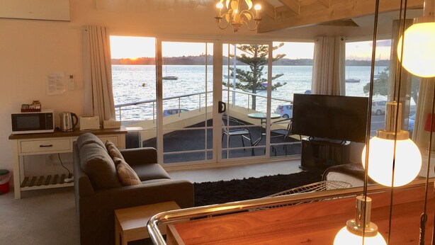 Un salón con sofá, televisor, mesa de centro de madera y vista al mar a través de amplios ventanales.
