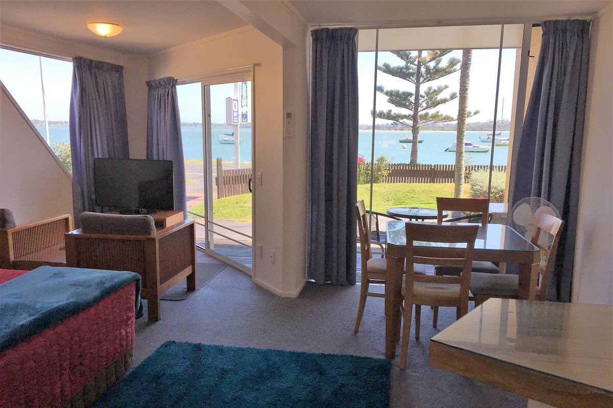 A room with a TV, a dining table, chairs, a sofa, and a view of the sea.