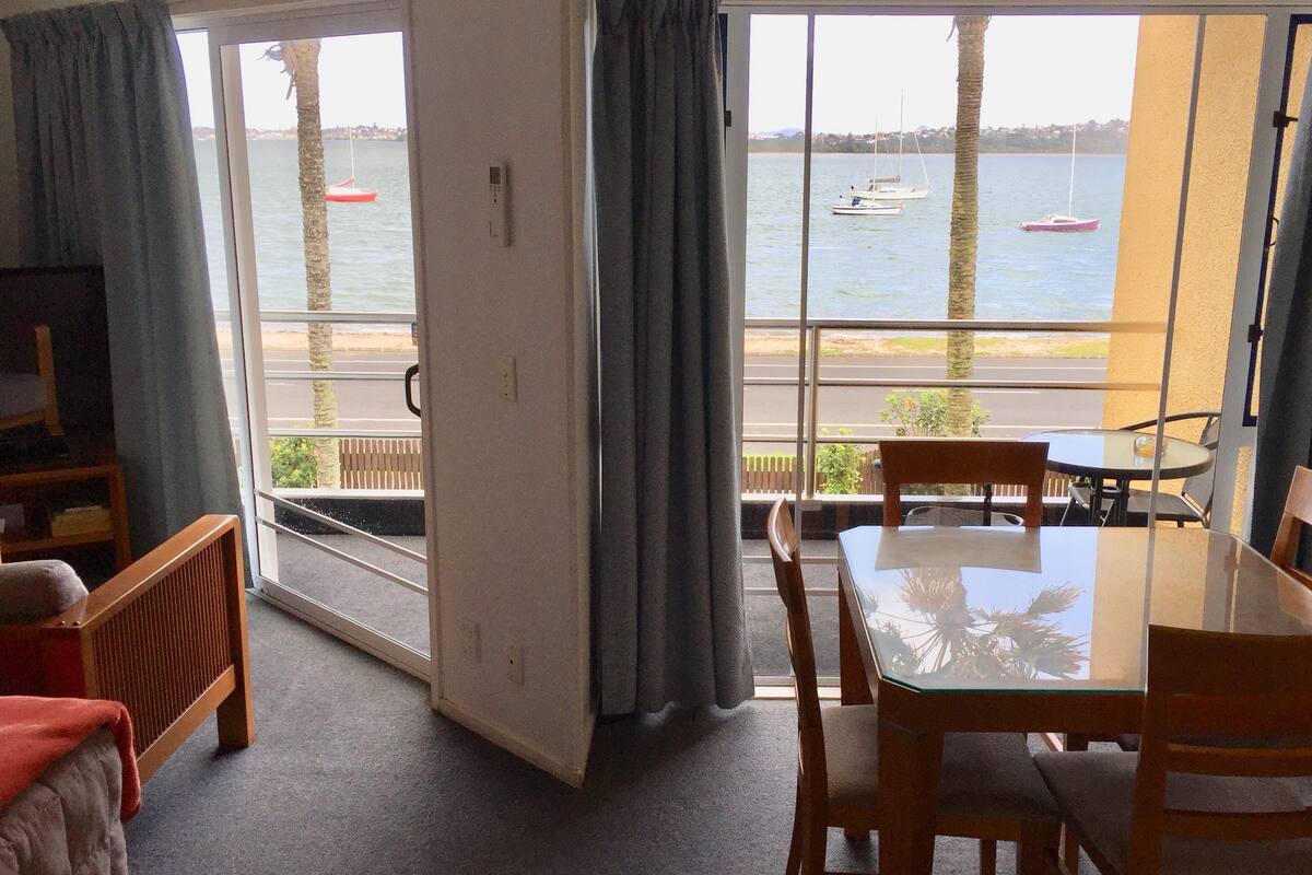 A room with a balcony overlooking a beach, a dining table, and a TV.