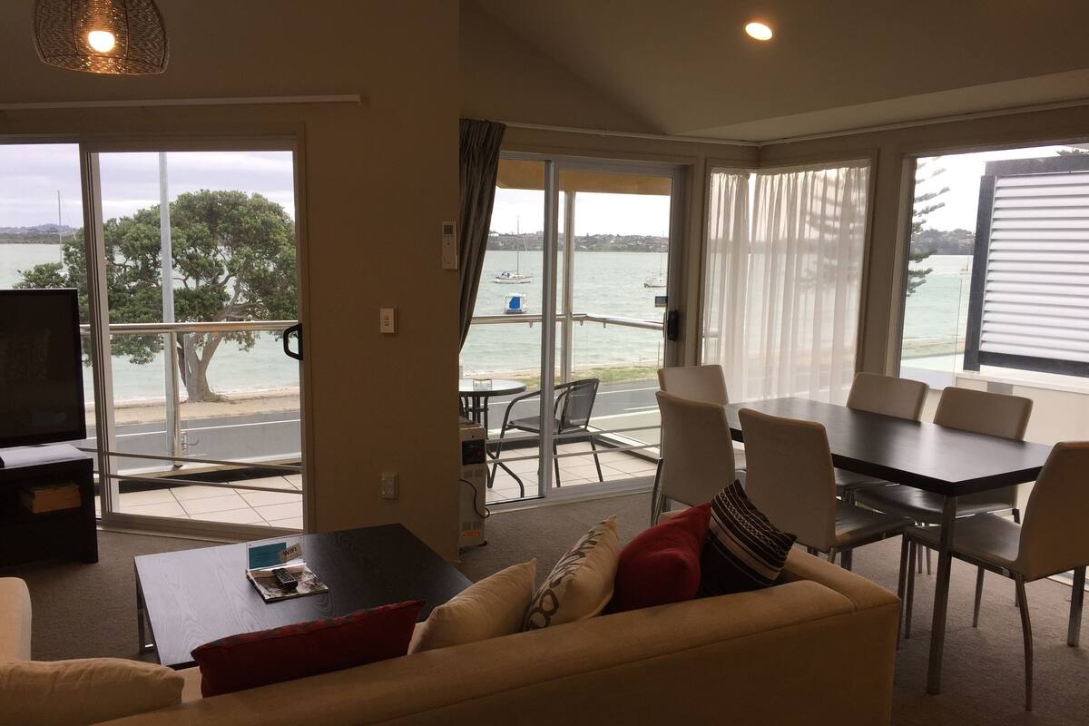 A living room with a sofa, a coffee table, and a TV. There is a balcony with a table and chairs, and a view of the sea.