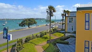 Una carretera costera con un edificio amarillo, palmeras y barcos al fondo.