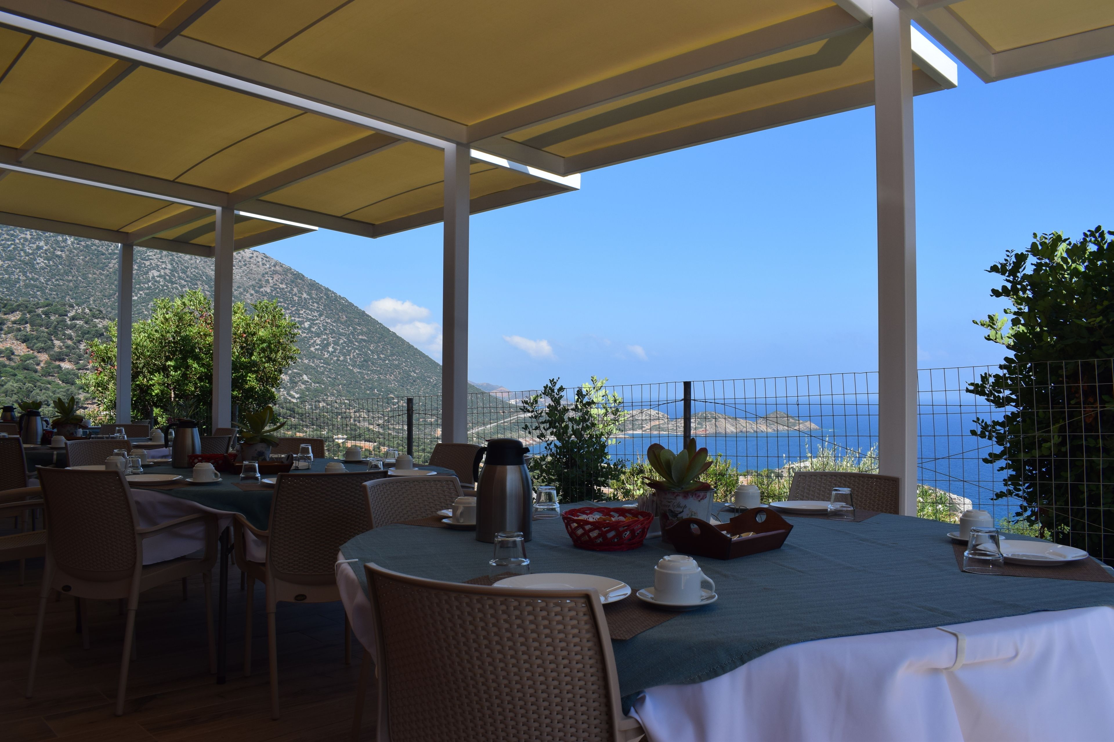 An outdoor dining area with tables set for two, overlooking a scenic view of the sea and mountains.