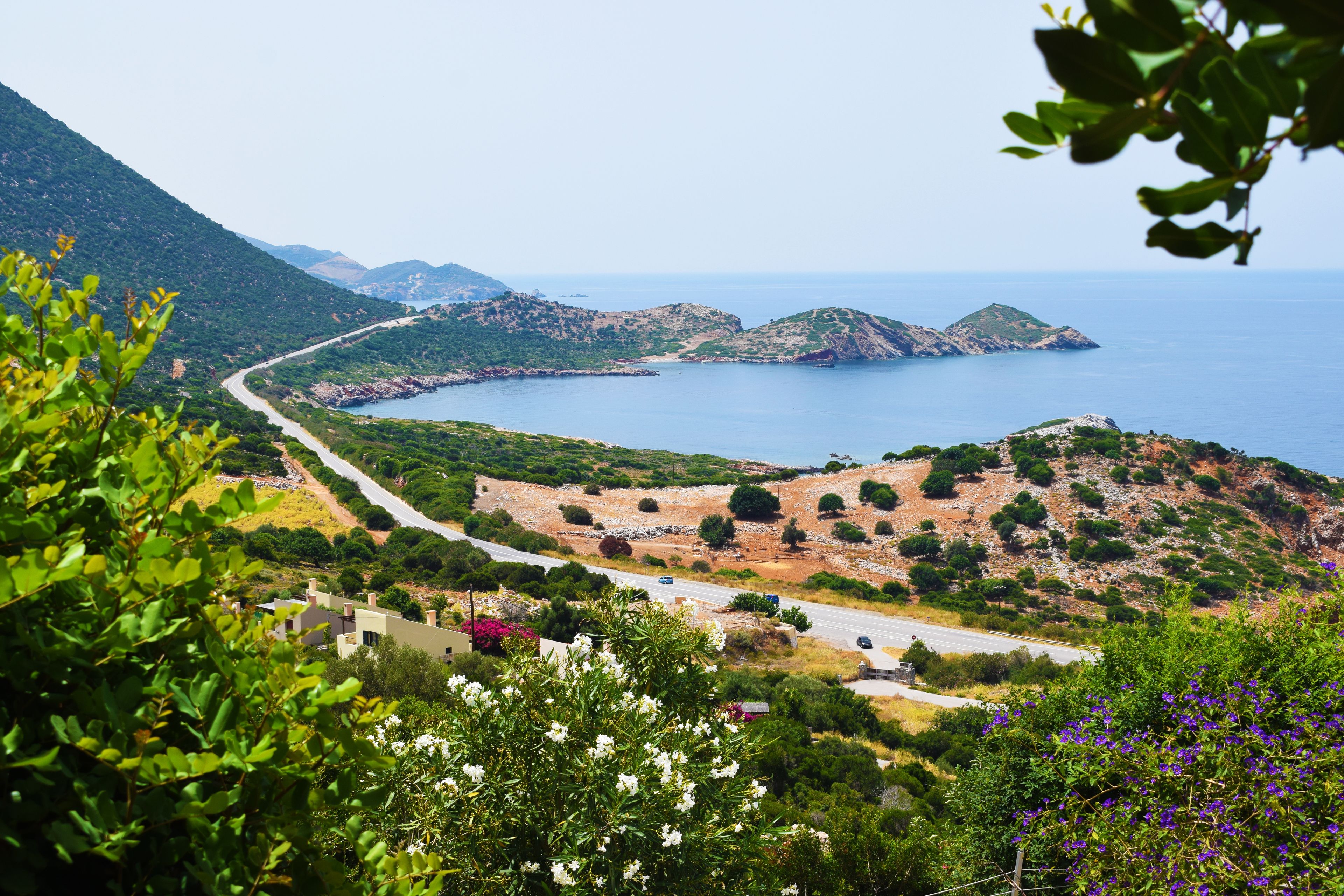 A coastal landscape with a winding road, green hills, and a clear view of the sea.