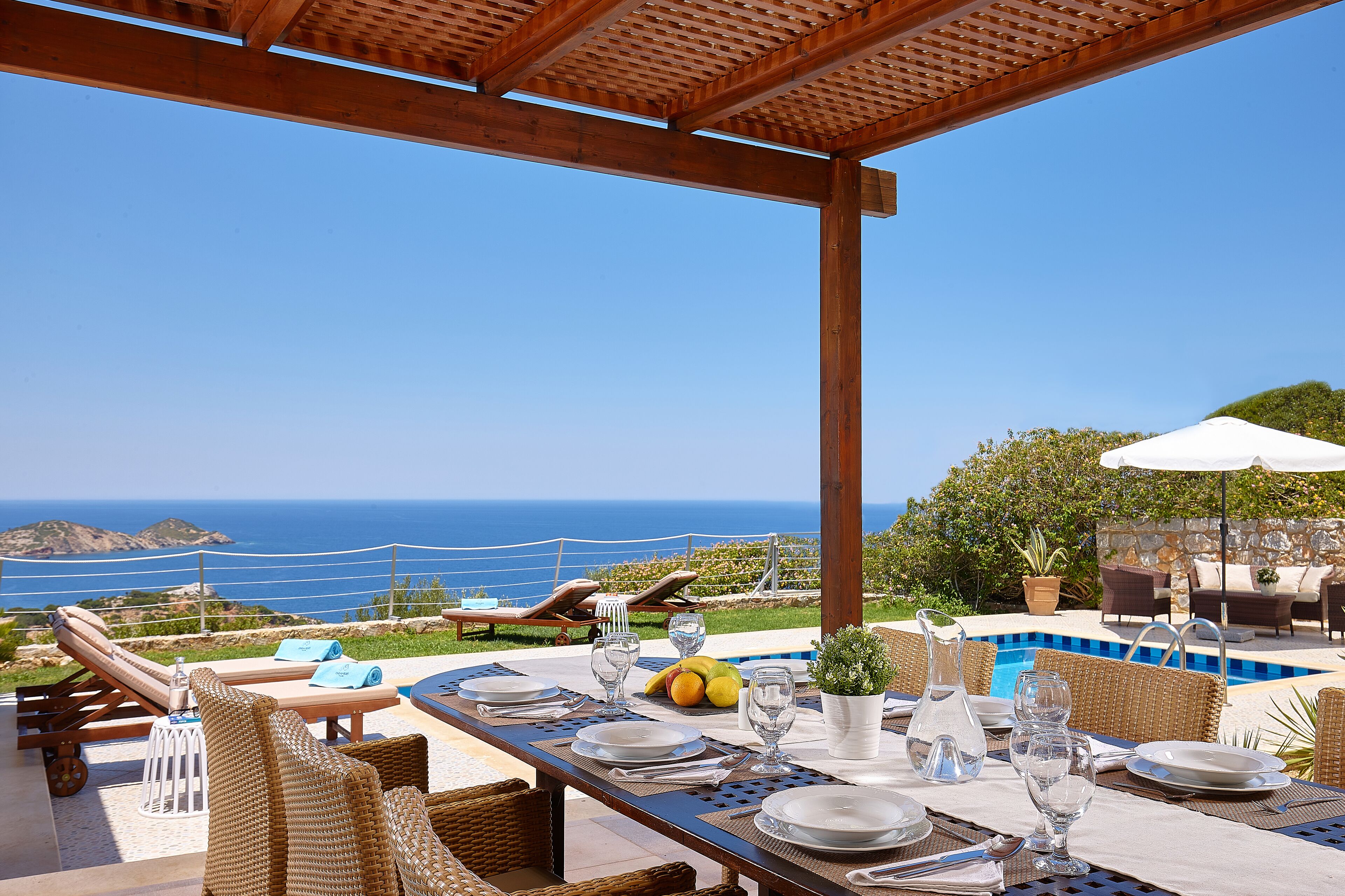 Un espacio de comedor al aire libre con vista al mar, equipado con mobiliario de mimbre, una piscina y una pérgola.