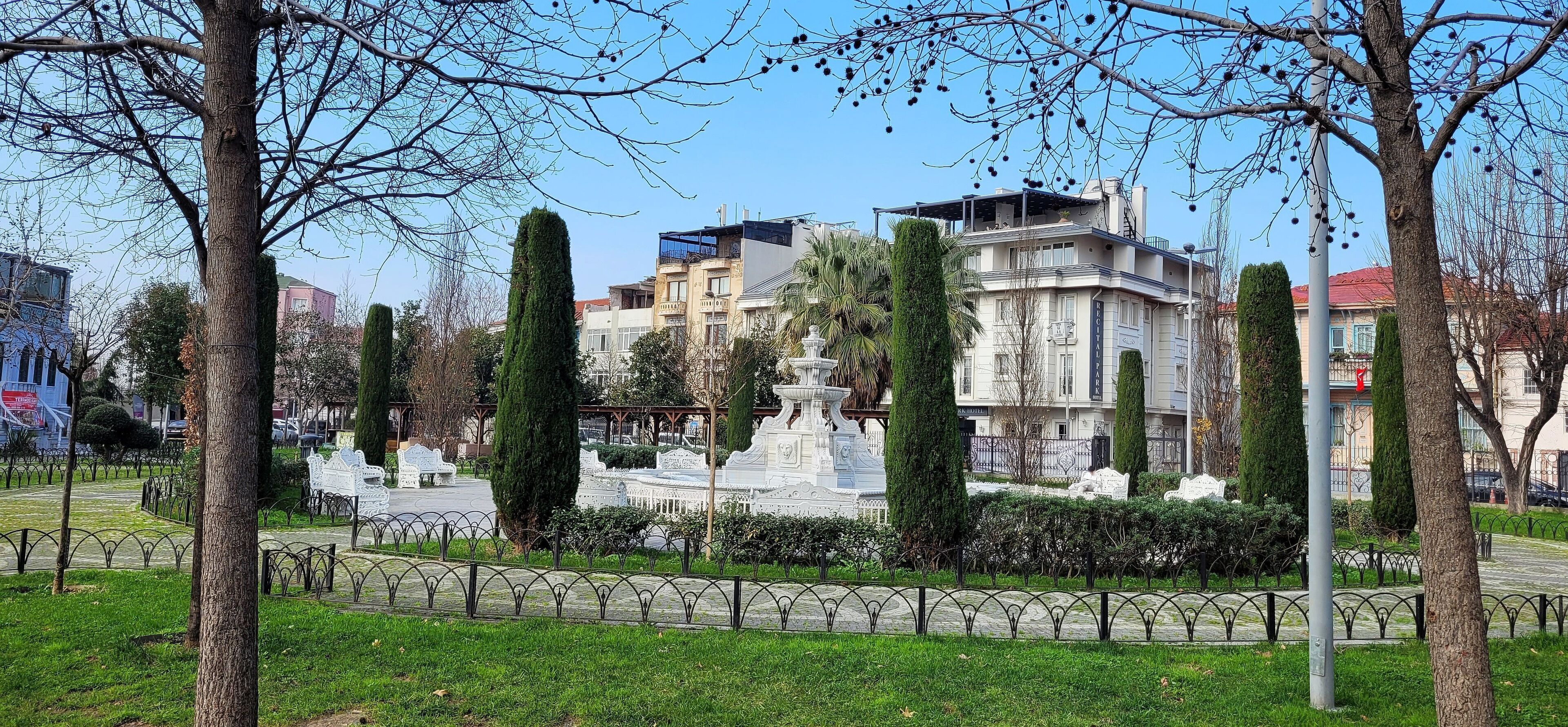 A park with a fountain, trees, and a building in the background.