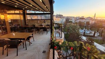 An outdoor seating area with tables and chairs, a view of the cityscape, and a decorative railing with greenery.